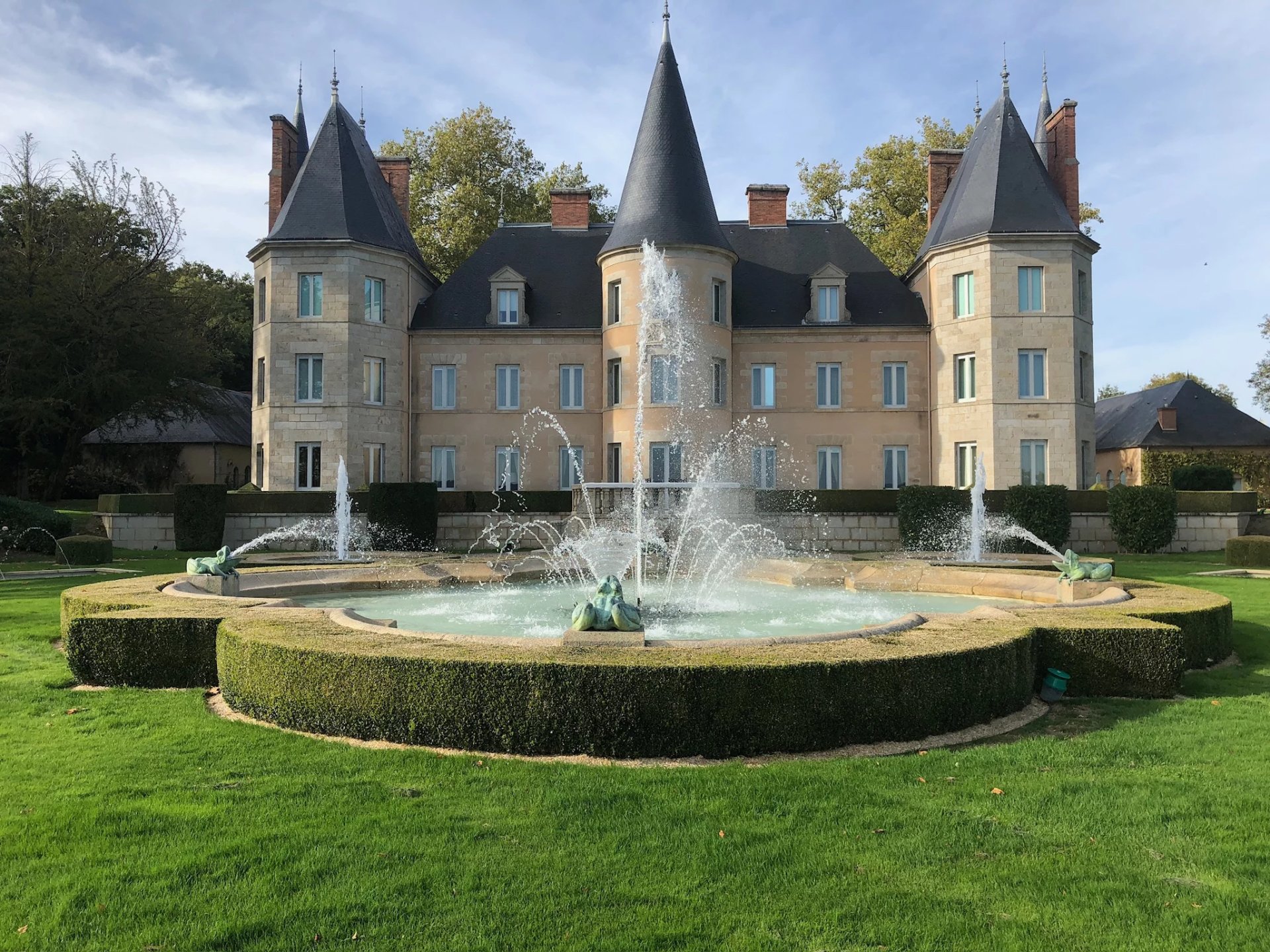 A fountain with statues shooting water in front of a chateau with roofs like castle spires.