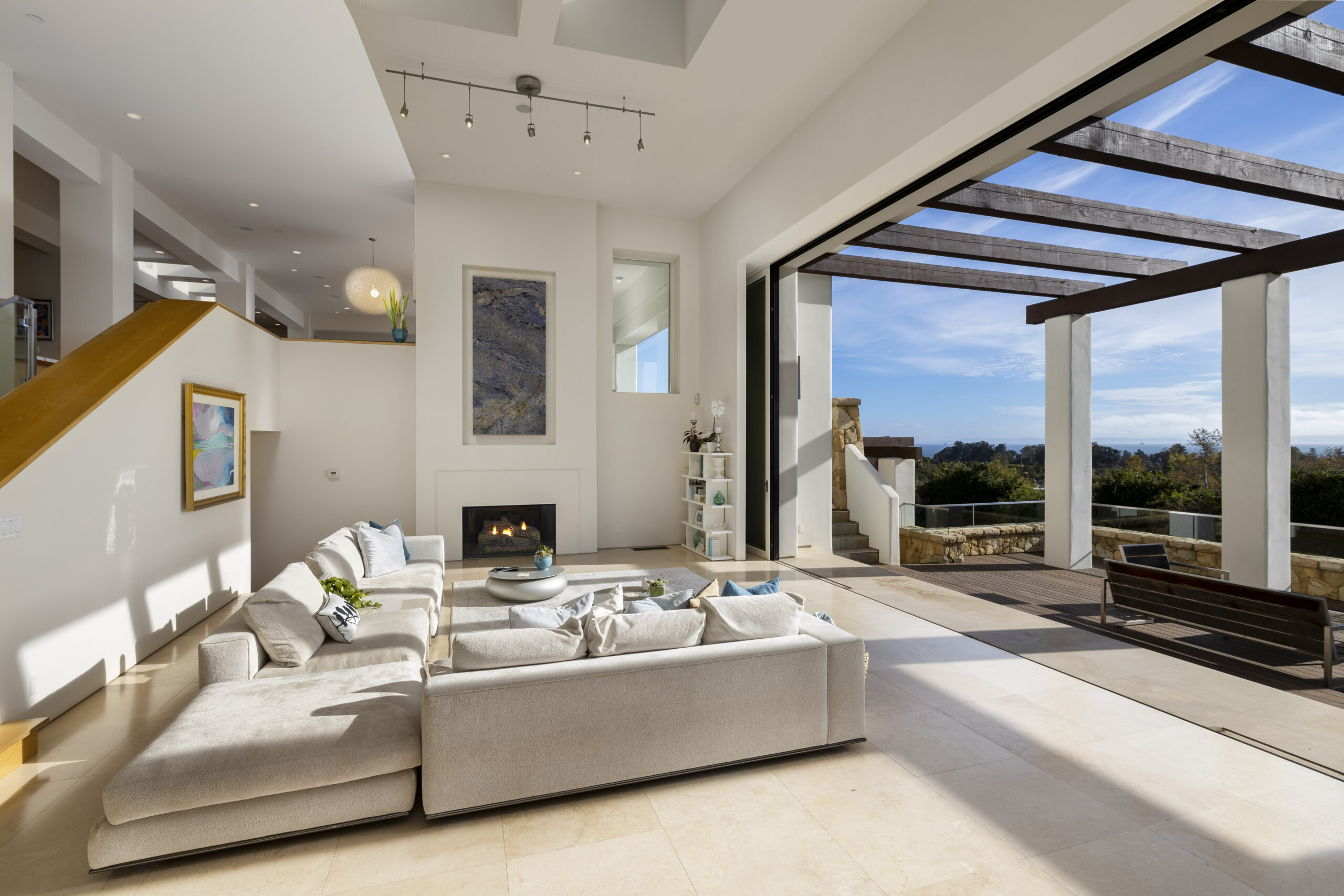 A pristine white living room with a panoramic window leading to a porch and blue skies.