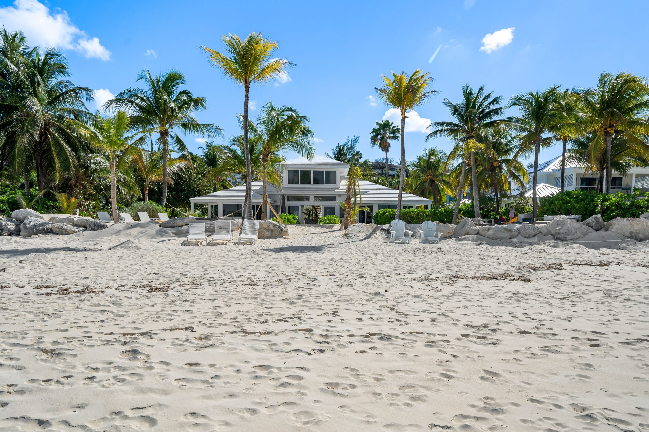 A narrow roofed resort home surrounded by palm trees in front of a clear beach.