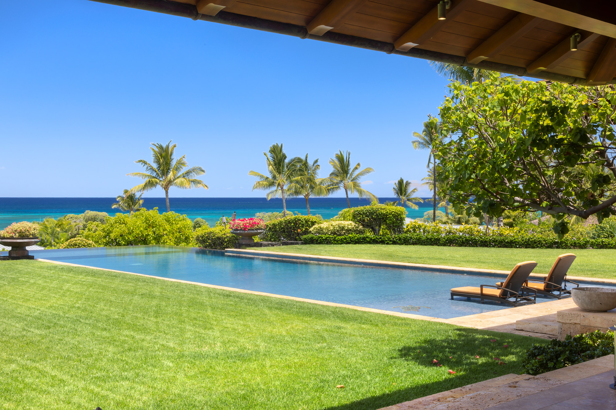 A long blue pool with pool chairs in it overlooking a green yard and blue skies.