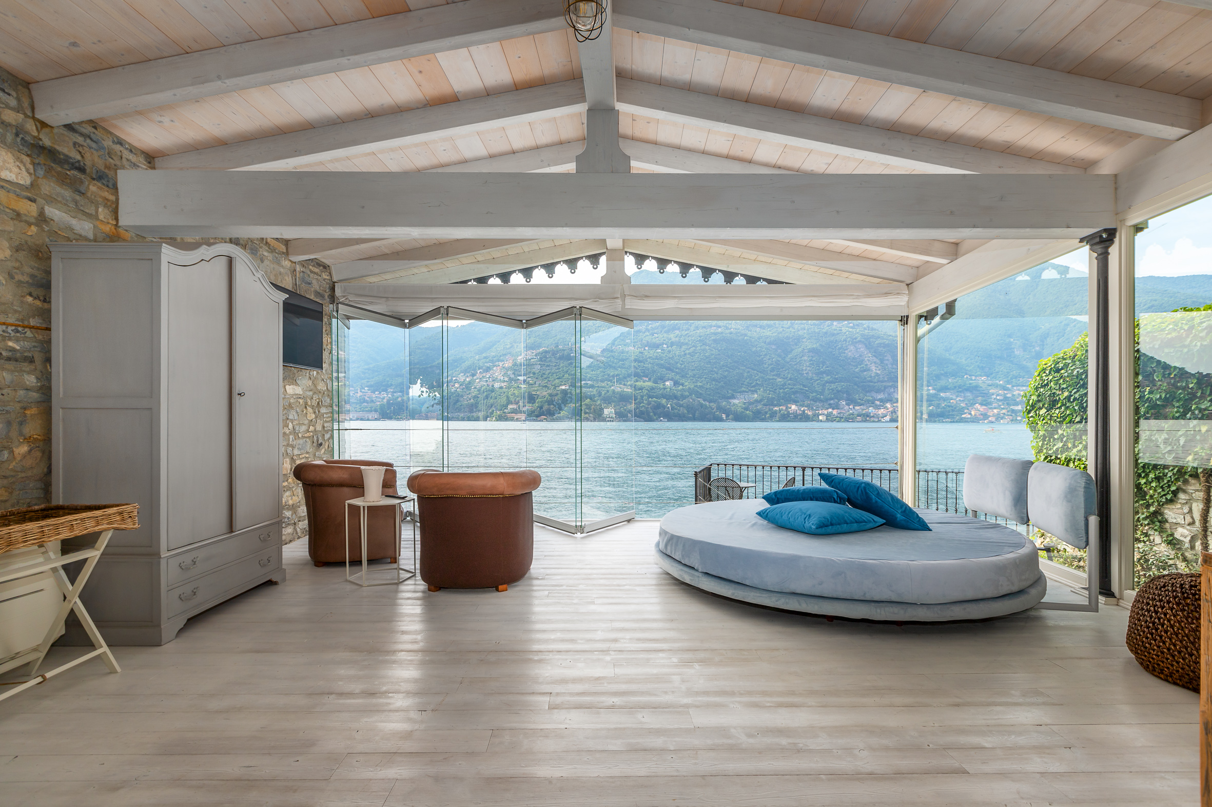 A room with a bed, dresser and a big glass wall with a nice view of the lake.