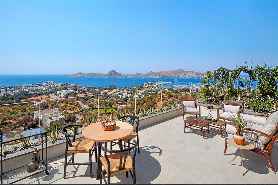 A balcony with a table and a sitting area that oversees a town below and a peninsula.