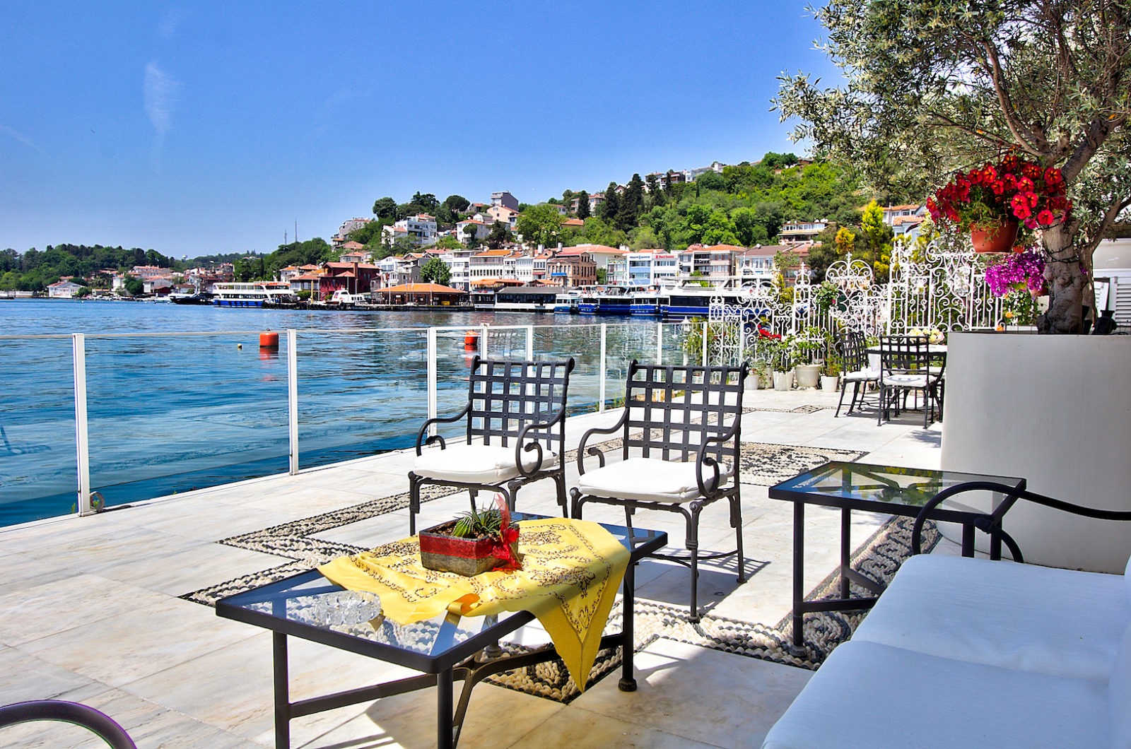 A porch with fancy chairs and flowers right next to the water with a town in the background.