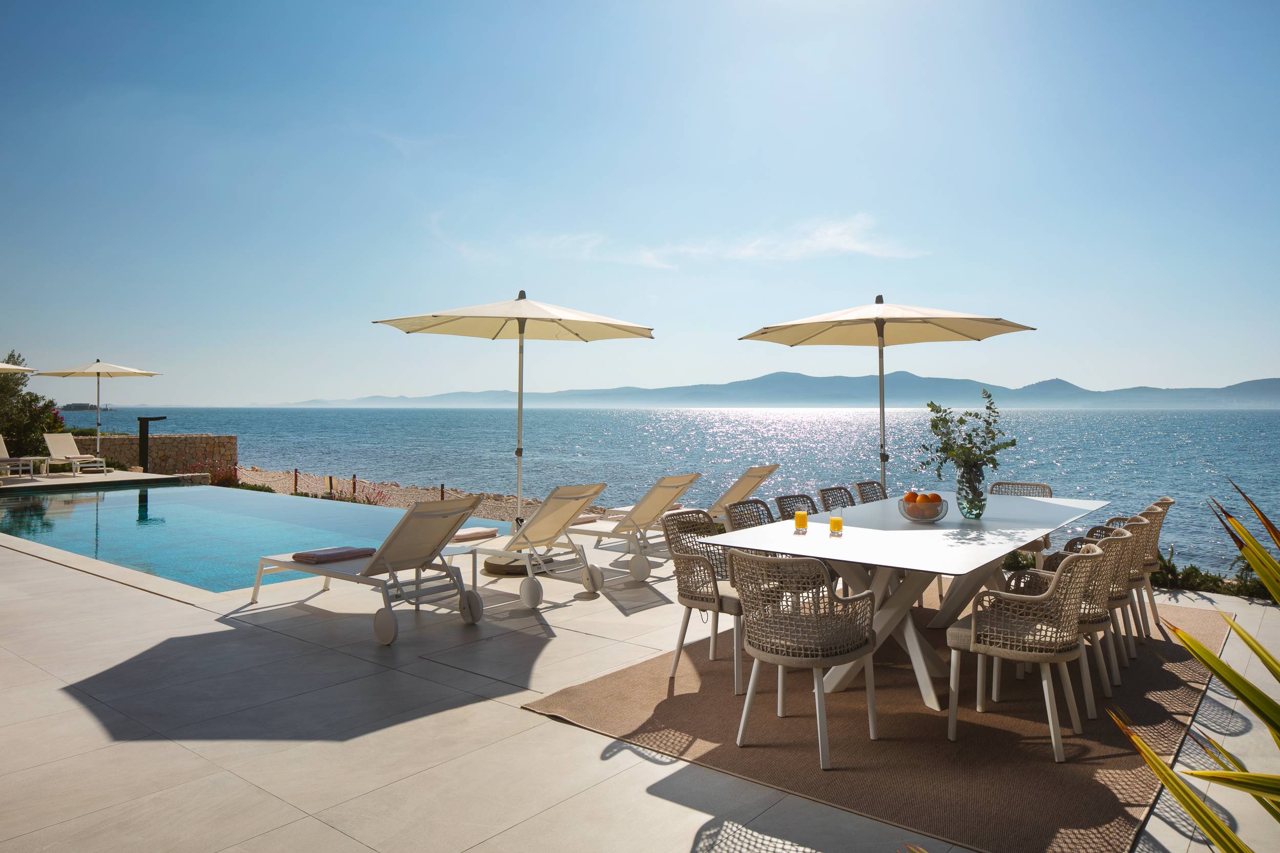 A pool next to tables and big umbrellas that oversees' a bay full of blue water.