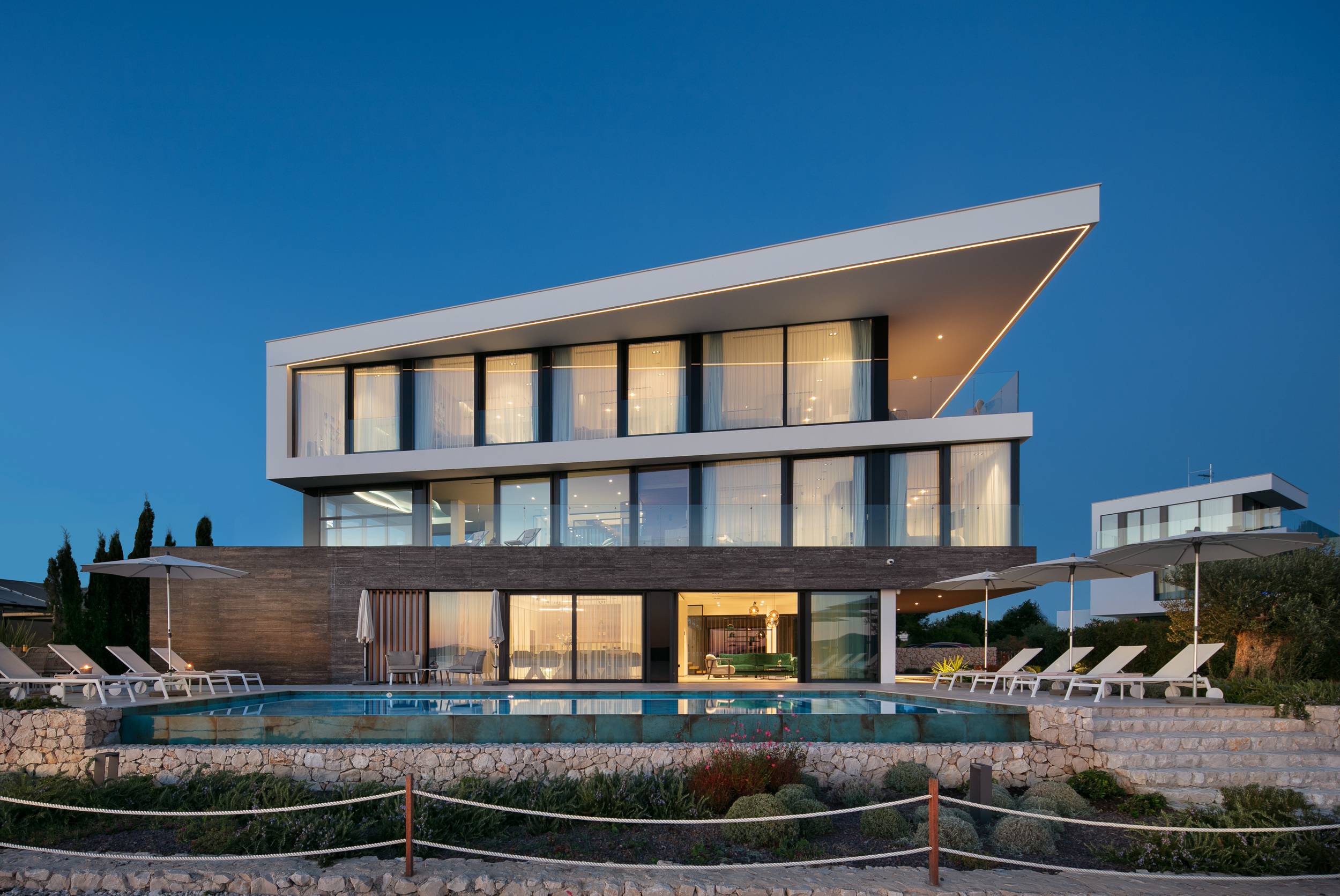 A house with glass walls and architecture that juts out at different angles. It has a pool and many poolside chairs.