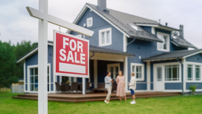 A For Sale sign in front of a house with a realtor and a young couple.