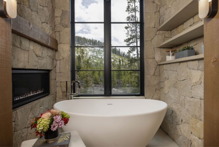 Soaking tub surrounded by limestone walls, a fireplace and window with mountain views. 
